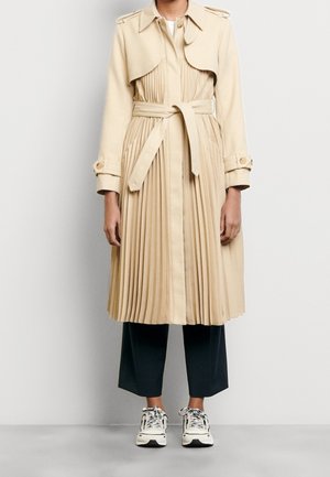 Beige veckad trench coat med midjeanpassad passform, bred krage och bälte; kombinerad med marinblå capribyxor och vita sneakers.