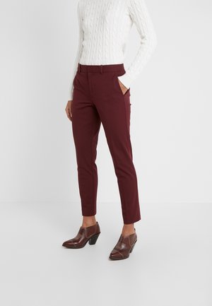 Personne portant un pantalon bordeaux ajusté aux chevilles, un pull blanc à torsades et des bottines marron à talons, debout avec une main dans la poche.