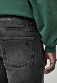 En person som bär svarta denimjeans och en grön långärmad topp, som visar bakfickan samt en del av ärmen och handen.