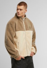 Jachetă din fleece în nuanțe de maron și crem, cu fermoar frontal, guler înalt, buzunare laterale și țesătură texturată. Include un patch cu logo pe piept.