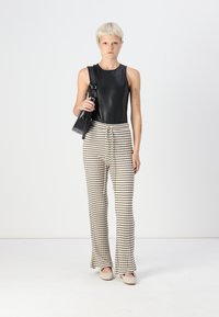 Svart ermeløs topp i faux skinn, parret med beige stripete bukser. Modellen holder en svart skulderveske og har på seg lyse slip-on sko.