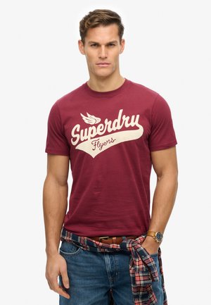En ung mand iført en mørkerød "Superdry Flyers" t-shirt, blå jeans, ternet skjorte bundet omkring livet og et armbåndsur, poserer foran en hvid baggrund.