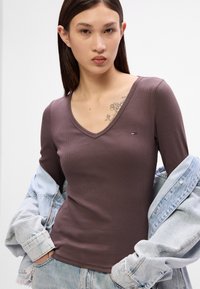 Top lung cu mânecă lungă, de culoare maro, cu decolteu în V. Prezintă un accent mic cu logo pe piept. Este asortat cu o jachetă din denim de culoare albastru deschis.