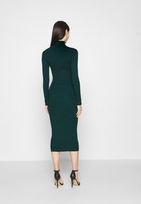 Robe en maille côtelée vert foncé avec col montant et manches longues, silhouette ajustée, longueur mi-mollet. Portée avec des chaussures à talons noires.