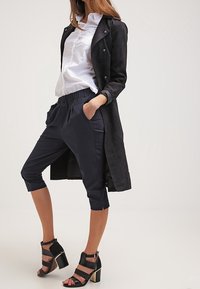 Manteau long en suede noir superposé à une chemise blanche boutonnée, associé à un pantalon cropped marine et des sandales à talons à lanières noires.