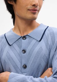 Pull en maille bleu clair avec un col classique, un passepoil bleu foncé, et cinq boutons texturés avec un motif en zigzag.