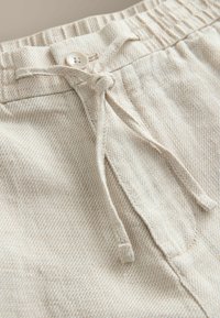 Gros plan d'un pantalon beige texturé avec un bouton, une ceinture élastique et des cordons en tissu noués à l'avant.