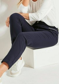 Marineblaue, taillierte Hose aus weichem Stoff, mit einer seitlichen Reißverschlusstasche, kombiniert mit weißen Turnschuhen und einem weißen Langarmoberteil.