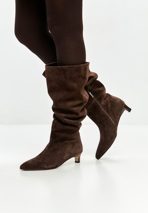 Bottes en daim marron, montantes, au design loose, avec un bout pointu et un petit talon, dotées d'une fermeture éclair sur le côté.