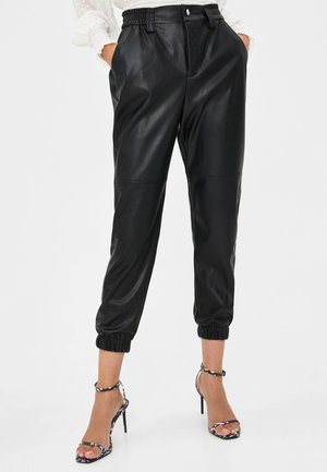 Zwarte joggingbroek van nepleer met elastische boorden en tailleband, gecombineerd met zwarte sandalen met hoge hak en bloemenmotief, handen in de zakken.