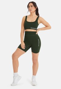 Top sportivo a coste verde scuro e pantaloni corti a vita alta abbinati, con la scritta "SQUATPROOF". La modella posa con scarpe da ginnastica bianche.