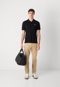 Zwart polo shirt met een klein wit logo, beige chino's, zwarte duffelbag en witte sneakers met beige accenten. Eenvoudig en functioneel ontwerp.