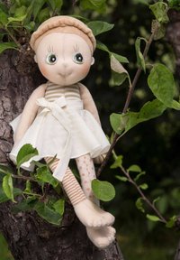 Miękka lalka z zielonymi oczami, w beżowej sukience z kokardą, w paskowane i kropkowane nogi, siedząca na gałęzi drzewa otoczona liśćmi.