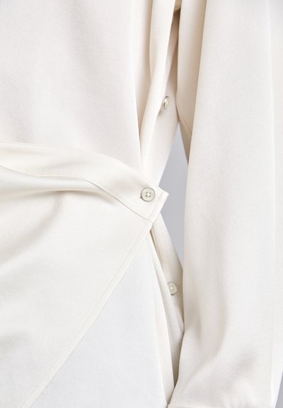 Blusa branca com uma textura suave, apresentando detalhes em botão e um design envolvente para um ajuste sob medida. O corte destaca a estrutura do tecido drapeado.