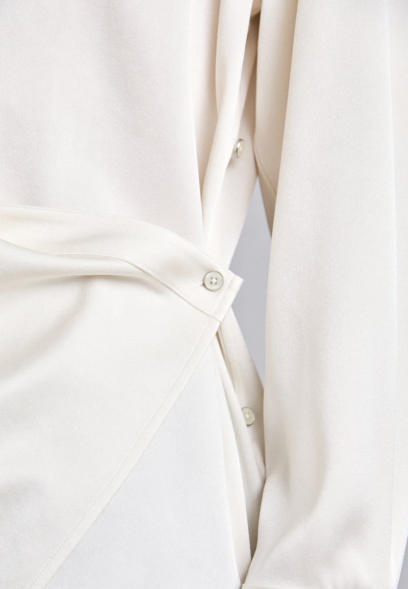 White blouse with a soft texture, featuring button details and a wrap design for a tailored fit. Cut showcases draped fabric structure.
