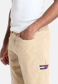 Pantaloni di cotone beige con una tasca sul lato sinistro, caratterizzati da una patch logo blu, bianca e rossa. Texture liscia, design aderente.