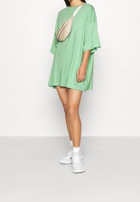 Vestido de camiseta verde de gran tamaño, hecho de algodón suave, con mangas cortas, combinado con un bolso cruzado beige y zapatillas blancas.