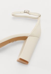 Scarpa beige con tacco a blocco quadrato, cinturino alla caviglia con fibbia metallica, materiale sintetico liscio e suola di un beige chiaro.