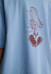 Illustration af en hånd med ben iført sneakers, der kører på et skateboard, trykt i brun på en lyseblå skjorte.