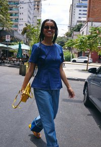Femme souriante aux cheveux tressés marchant dans la rue de la ville, portant un haut bleu, un jean, des sandales jaunes, des lunettes de soleil et un sac à main jaune.