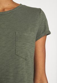 Oliwkowy t-shirt z krótkim rękawem, wykonany z miękkiej, teksturowanej tkaniny, z kieszonką na piersi i okrągłym dekoltem.