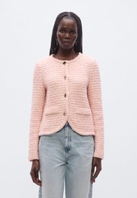 Rózsaszín texturált dzseki kerek nyakkivágással, arany gombokkal és két első zsebbel. Világoskék farmernadrággal párosítva.