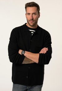 Cardigan nera a bottoni con colletto, tessuto a trama, e taschino sul petto, abbinata a una camicia a righe sotto; indossata con jeans.