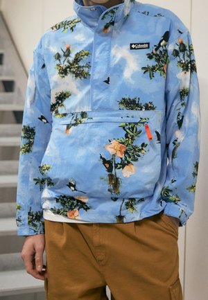 Mężczyzna w jasnoniebieskiej bluzie Columbia z zielonym wzorem kwiatów i ptaków, w brązowych spodniach, stojący wewnątrz blisko schodów.