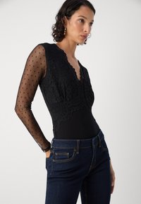 Body negro con detalles de encaje y mangas de lunares transparentes, combinado con jeans de mezclilla oscuros. Cuello notable con forma de scalloped y acentos de costura.