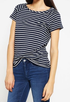 Kobieta w granatowo-białej koszulce w paski z krótkim rękawem, lekko włożonej do niebieskich dżinsów, stojąca na jednolitym tle.