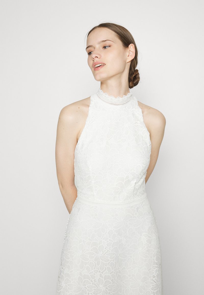 Vestido blanco de encaje con un alto escote festoneado y un patrón floral. Cuerpo ajustado que fluye hacia una falda amplia, con una textura delicada.