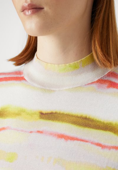 Strikkegenser med ribbestrikket krage, med et flerfarget, abstrakt mønster i gul, rosa og brun på hvit bakgrunn.
