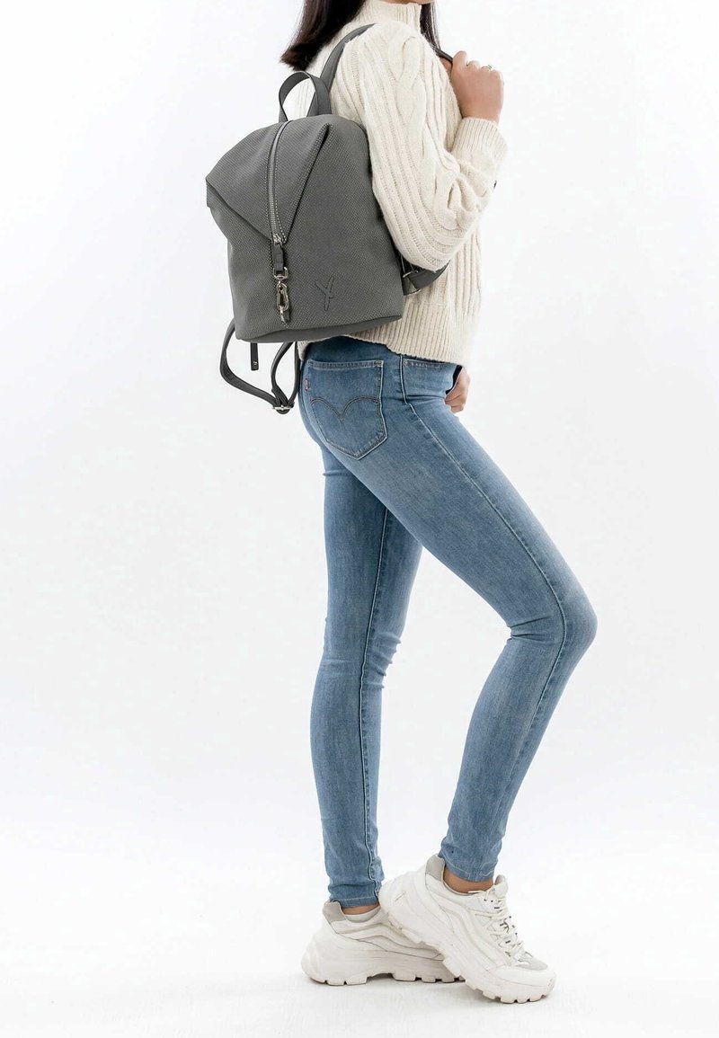 Personne portant un jean skinny bleu, des baskets blanches épaisses, un pull en maille torsadée crème, et portant un sac à dos gris texturé sur une épaule.