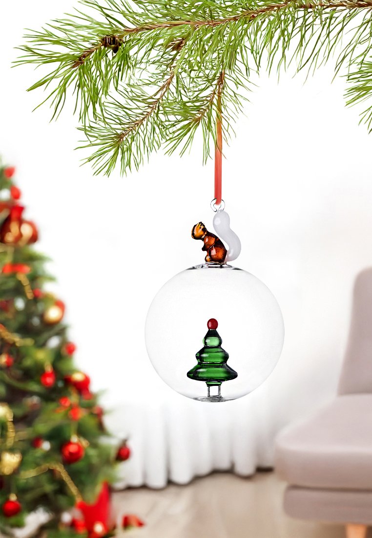 Przezroczysta szklana ozdoba choinkowa z zielonym wzorem drzewa, z czerwonym akcentem, zwieńczona brązową wiewiórką, zawieszona na czerwonej wstążce.
