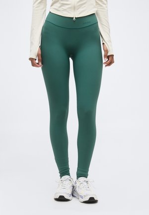 Άτομο που φοράει κολάν υψηλής μέσης σε τιρκουάζ απόχρωση, κρεμ μακρυμάνικη μπλούζα με τρύπες για τους αντίχειρες και λευκά αθλητικά παπούτσια, σε απλό λευκό φόντο.