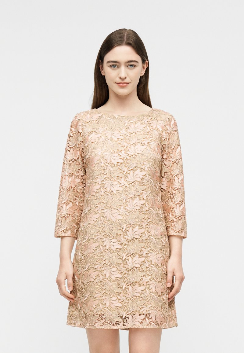 Νέα γυναίκα με μακριά καστανά μαλλιά, που φοράει ένα ροζ δαντελένιο φόρεμα με λουλούδια και μανίκια τριών τετάρτων, στέκεται μπροστά σε ένα απλό φωτεινό φόντο.