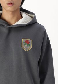 Szara bluza z kapturem z białą teksturowaną podszewką, z haftowaną naszywką przedstawiającą czerwoną różę i zielone liście, oprawioną w złoto.