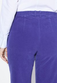 Paarse corduroy broek met een elastische tailleband, voorzien van platte achterzakken en een gestructureerde afwerking. Het model draagt een wit shirt met lange mouwen.