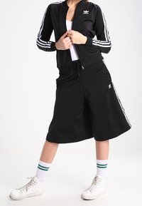 Černá sportovní bunda s bílými pruhy, odpovídající černé kraťasy a bílé tenisky se zelenými akcenty. Hladká tkanina, každodenní sportovní design.