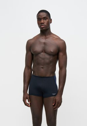 Veltrænet mand står mod en hvid baggrund iført sorte Speedo badetrusser, kigger lige frem med afslappet kropsholdning.