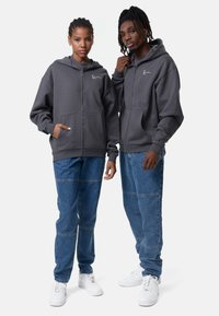 Grå zip-up hættetrøjer med frontlommer, båret med blå denim jeans. Bukserne har stikninger, og begge bærer hvide sneakers.