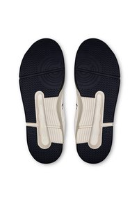 Des baskets blanches avec des semelles en caoutchouc noir, présentant un motif de grip texturé et un accent en plastique au niveau du talon. Le design est minimaliste et élégant.