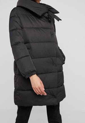 Person trägt einen schwarzen, gesteppter Steppmantel mit elastischen Bündchen, großem Kragen mit Bindebändern und Seitentaschen vor einfachem Hintergrund.