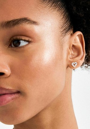 Pendiente de botón plateado en forma de corazón con centro abierto, llevado en la oreja izquierda. Textura metálica lisa, vista en primer plano junto al tono de piel.