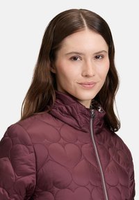 Donna con lunghi capelli castani che indossa una giacca trapuntata bordeaux con motivi a cuore e un colletto con zip, rivolta leggermente a destra con un'espressione neutra.