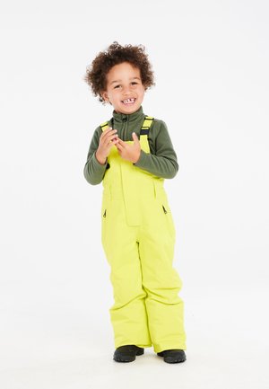 Gelbe wasserdichte Latzhose mit verstellbaren Trägern und Taschen, kombiniert mit einem grünen Fleeceoberteil. Kind steht lächelnd vor einem weißen Hintergrund.