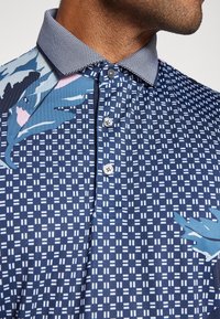 Polo azul con estampado de diseño geométrico, cuello en contraste y botones a lo largo de la tapeta. Textura suave y tela ligera.