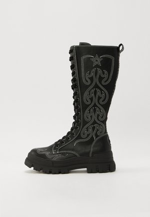 Botine din piele neagră, cu șiret, având un ștaif înalt, decorate cu cusături albe, modele de flacără și o talpă groasă, texturată.