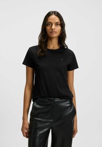 Schwarzes Baumwoll-T-Shirt mit Rundhalsausschnitt und kurzen Ärmeln, das ein dezentes Logo auf der Vorderseite trägt; kombiniert mit glänzenden schwarzen Hosen.