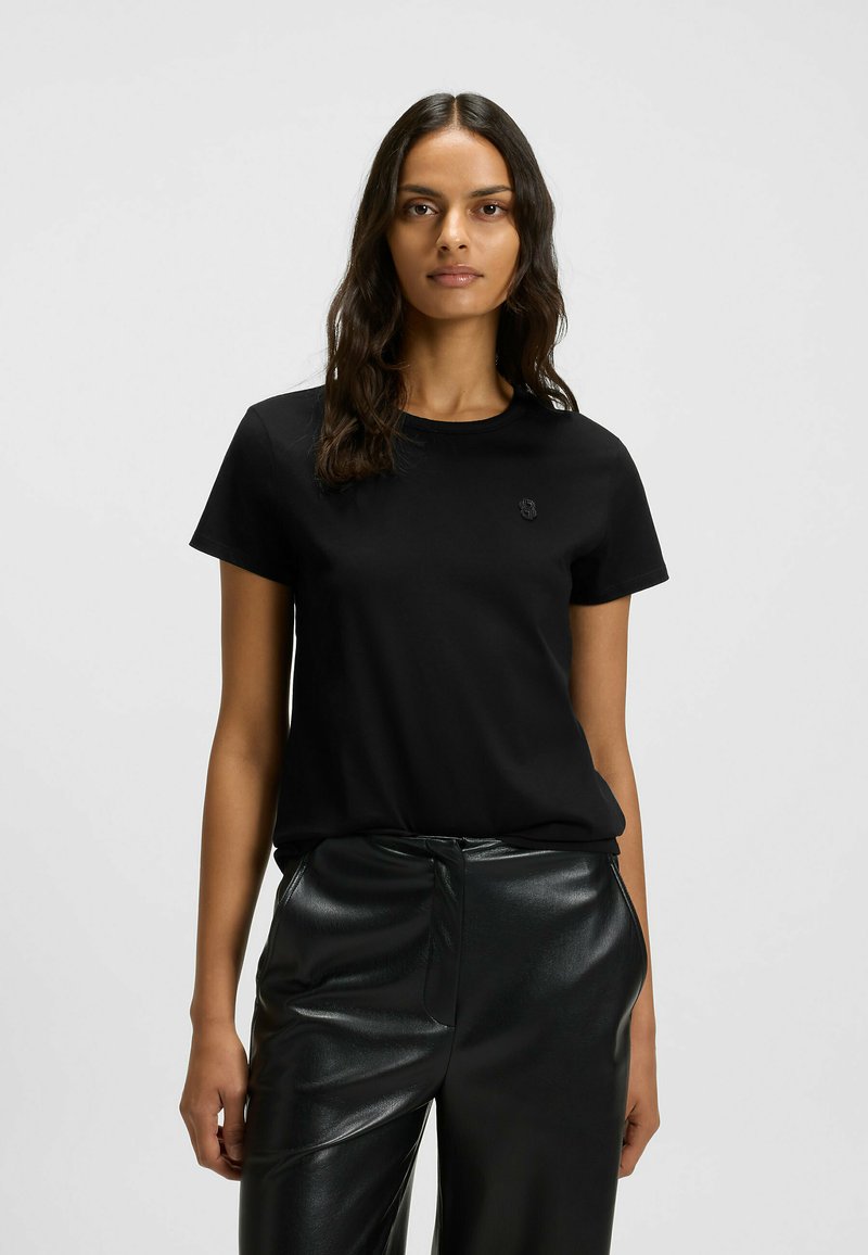 Schwarzes Baumwoll-T-Shirt mit Rundhalsausschnitt und kurzen Ärmeln, das ein dezentes Logo auf der Vorderseite trägt; kombiniert mit glänzenden schwarzen Hosen.